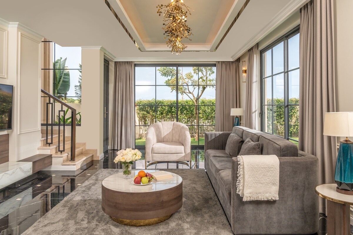 A spacious living area is depicted, featuring modern furnishings and large windows that allow natural light. A plush gray sofa faces a round coffee table adorned with fresh fruit and flowers. Elegant light fixtures complement the high ceiling, while decorative curtains frame the view.