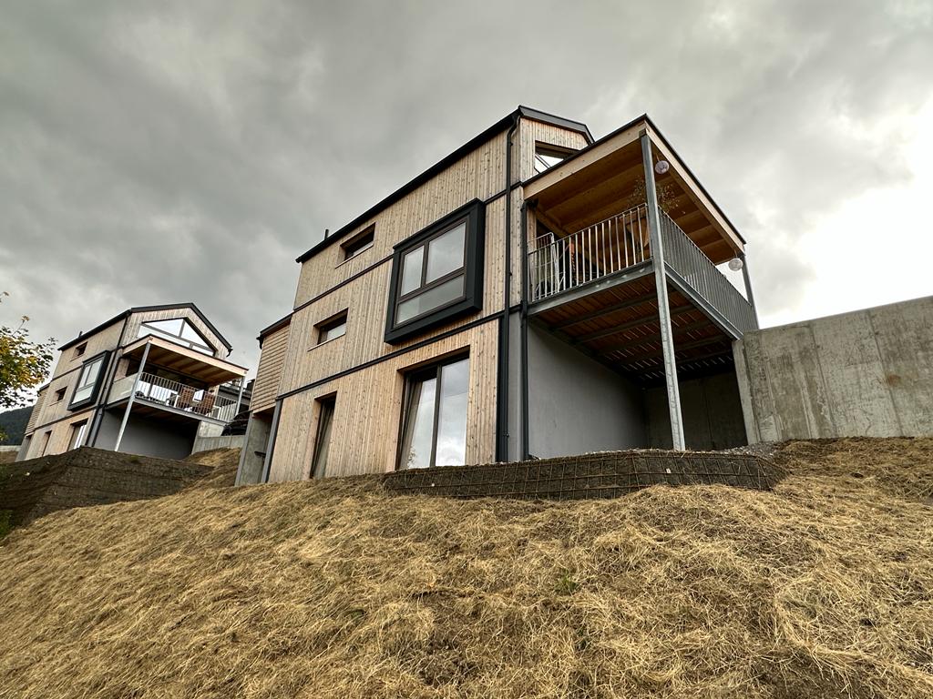 The modern two-story tiny house is framed by a sloping landscape, featuring a combination of wood and concrete finishes. Large windows enhance the structure's facade, and a balcony extends from the upper level, overlooking the natural surroundings beneath a cloudy sky.
