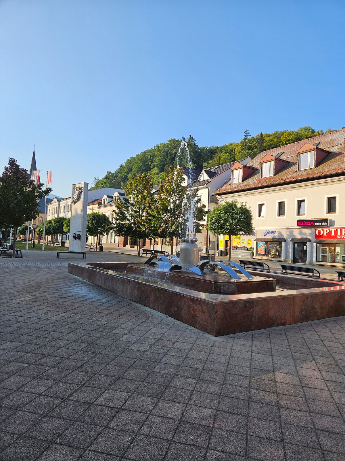 A public square is presented, featuring a central water fountain with cascading water. Surrounding greenery includes trees and shrubs. Nearby buildings exhibit traditional architecture, while benches provide seating along the plaza. A clear blue sky completes the scene, adding to the area's inviting atmosphere.