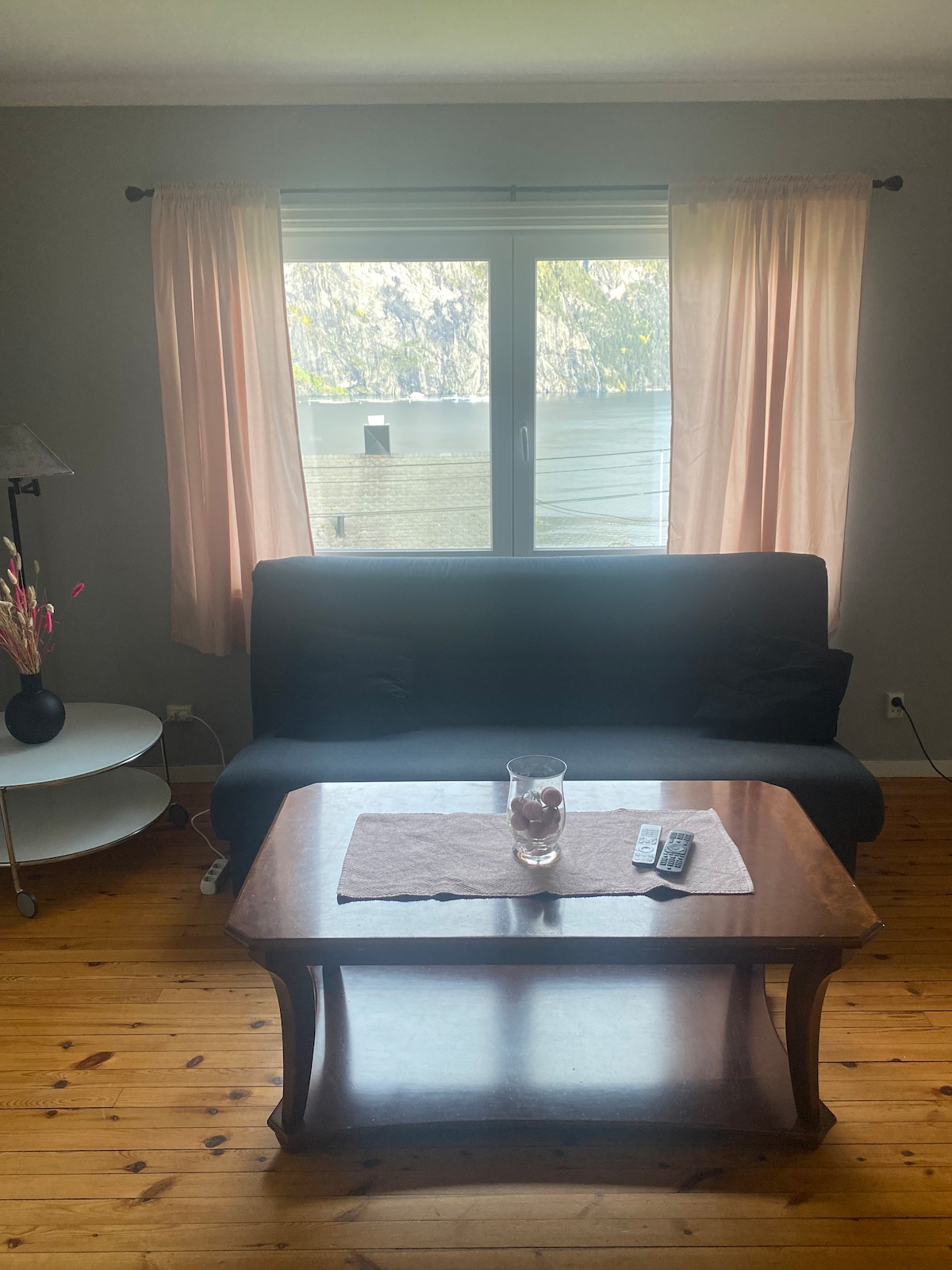 A cozy living area features a dark sofa positioned in front of a window, allowing natural light to enter the space. A wooden coffee table is centered beneath the window, topped with a decorative item and a remote control. Soft curtains frame the window.