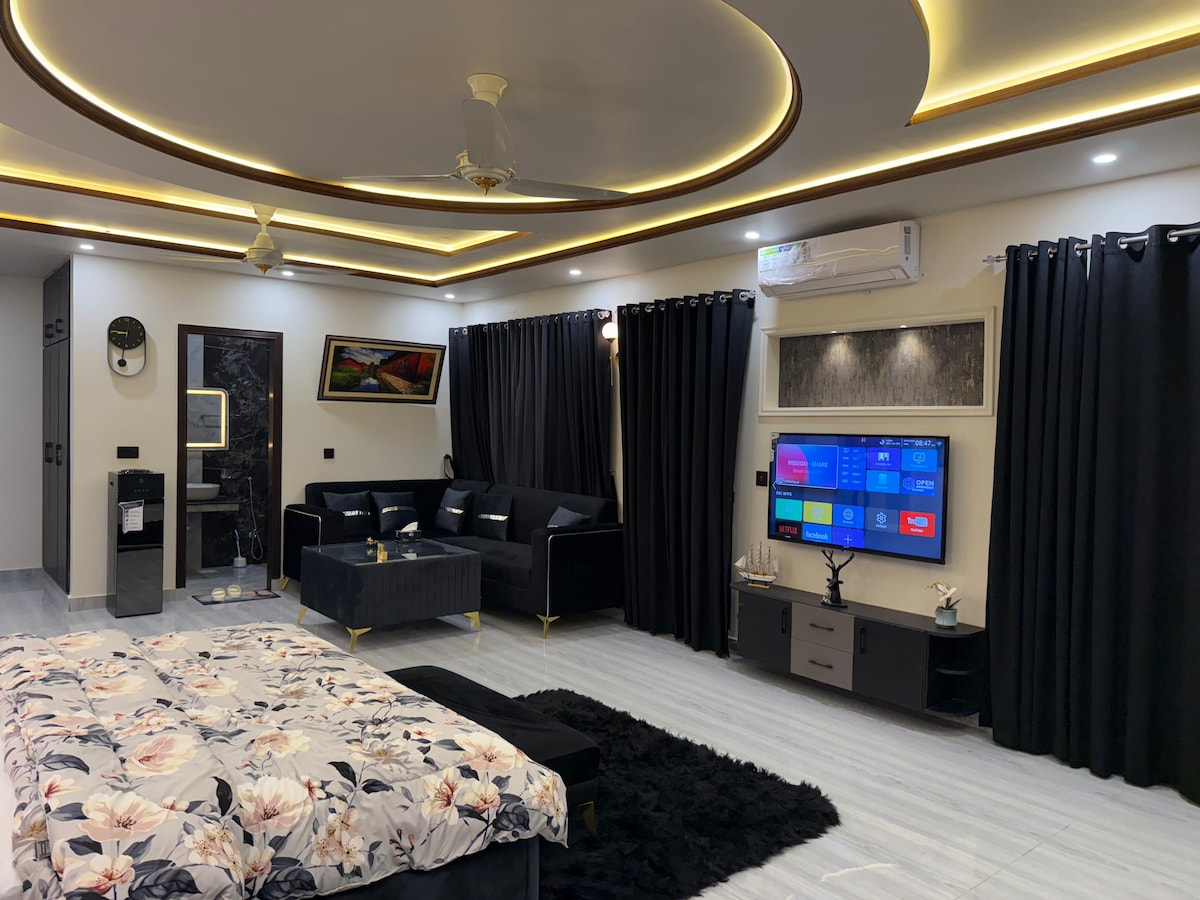 A spacious living area features a large floral-patterned bedspread, complemented by a contemporary black sofa set. Dark curtains frame the windows, and a flat-screen TV is mounted on the wall. Ambient lighting enhances the modern decor, creating a cozy and inviting atmosphere.