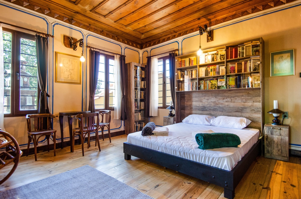 A bright bedroom features a spacious bed with a wooden headboard and an assortment of decorative items. Natural light floods through multiple windows with curtains. A small dining table is situated near a bookshelf, creating an inviting reading and dining area.