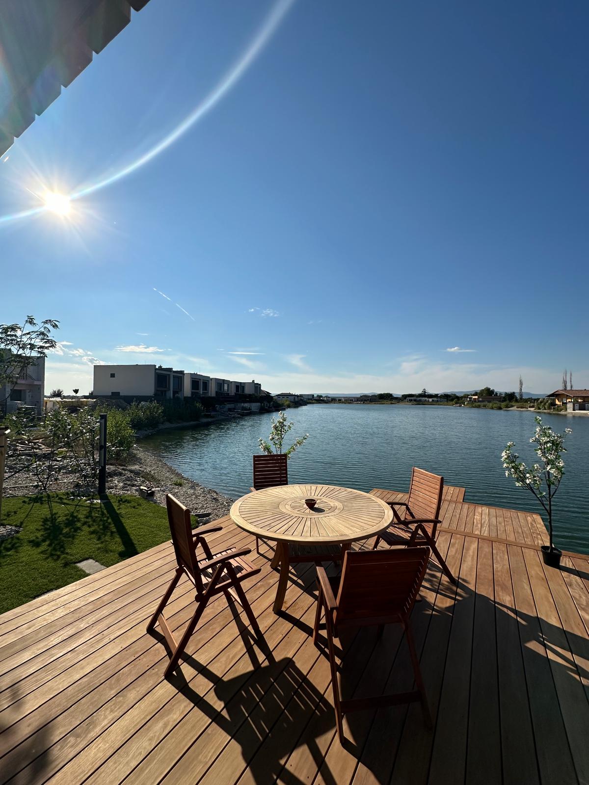 A wooden deck overlooking a calm lake features a round table surrounded by five wooden chairs. Sunlight reflects off the water, while gentle foliage adds a natural touch to the serene setting. Modern mobile homes are visible in the background against a clear blue sky.