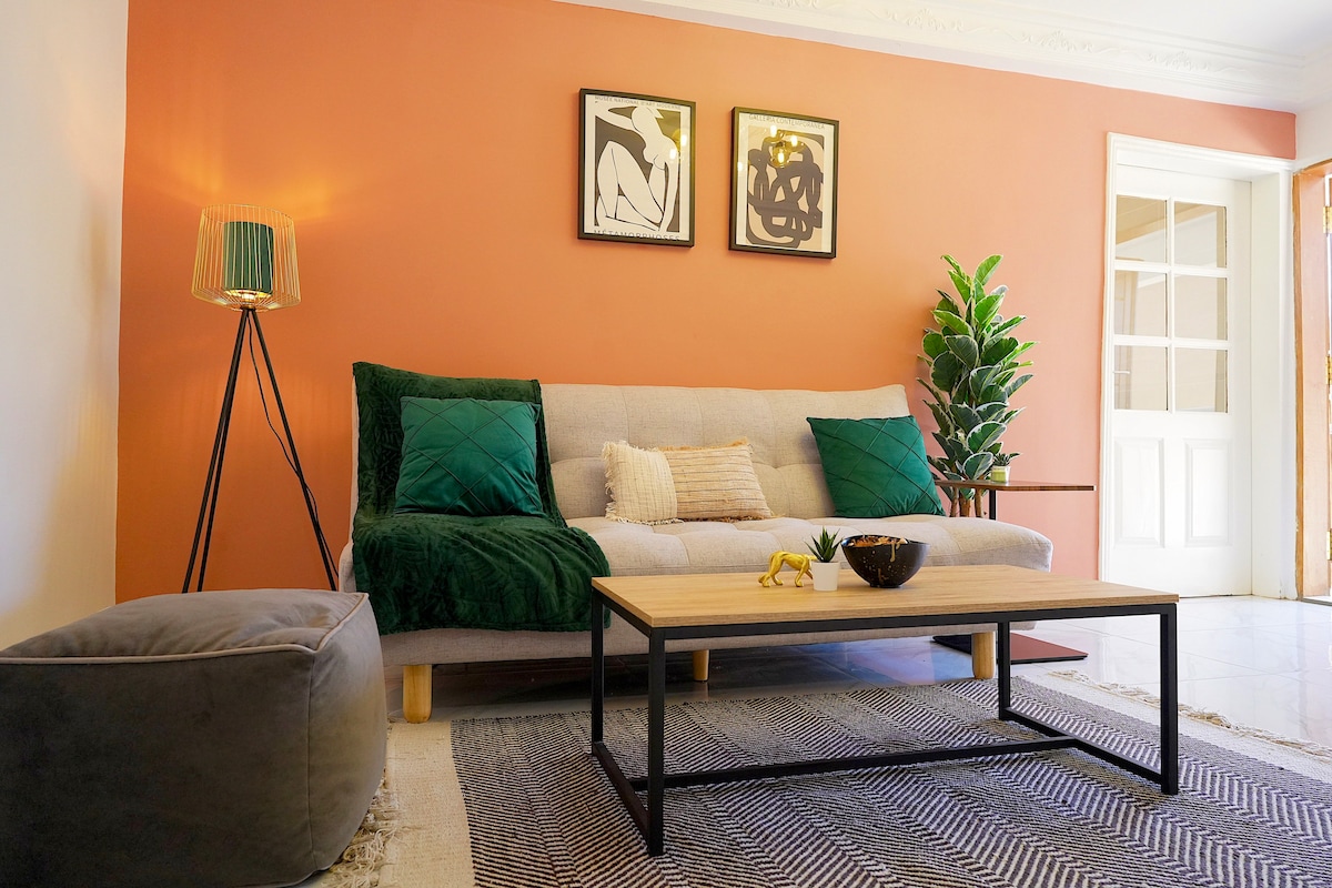 A living area features a light-colored sofa adorned with green cushions, complemented by a wooden coffee table and a textured area rug. Green plants add a natural touch, while framed artworks adorn the orange wall, creating a cozy and well-lit environment.