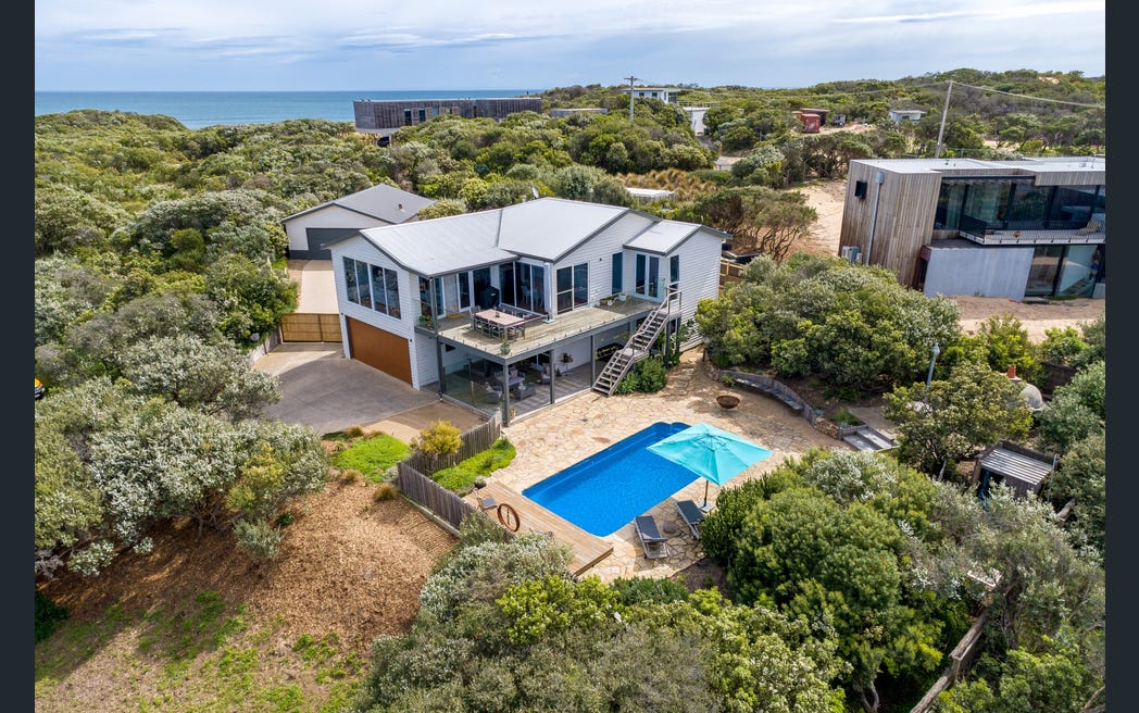 An aerial view of a modern family home surrounded by lush greenery and sandy terrain, with the ocean visible in the background. A spacious outdoor area features a solar-heated pool and various seating arrangements on a stone patio, providing an inviting space for relaxation.