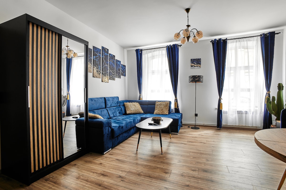 A stylish living area features a blue sectional sofa complemented by decorative cushions. Natural light is filtered through tall windows adorned with dark curtains. A modern coffee table is placed centrally, while a mirror-fronted cabinet enhances the room's depth. Wall art adds a contemporary touch.