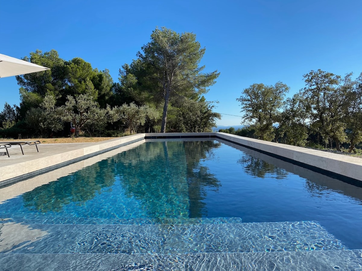 A serene salt-water pool is shown, featuring a shallow entry point. The clear water reflects the surrounding trees and blue sky, creating a calming effect. The pool area includes a spacious deck, offering views of the countryside and ample space for relaxation.