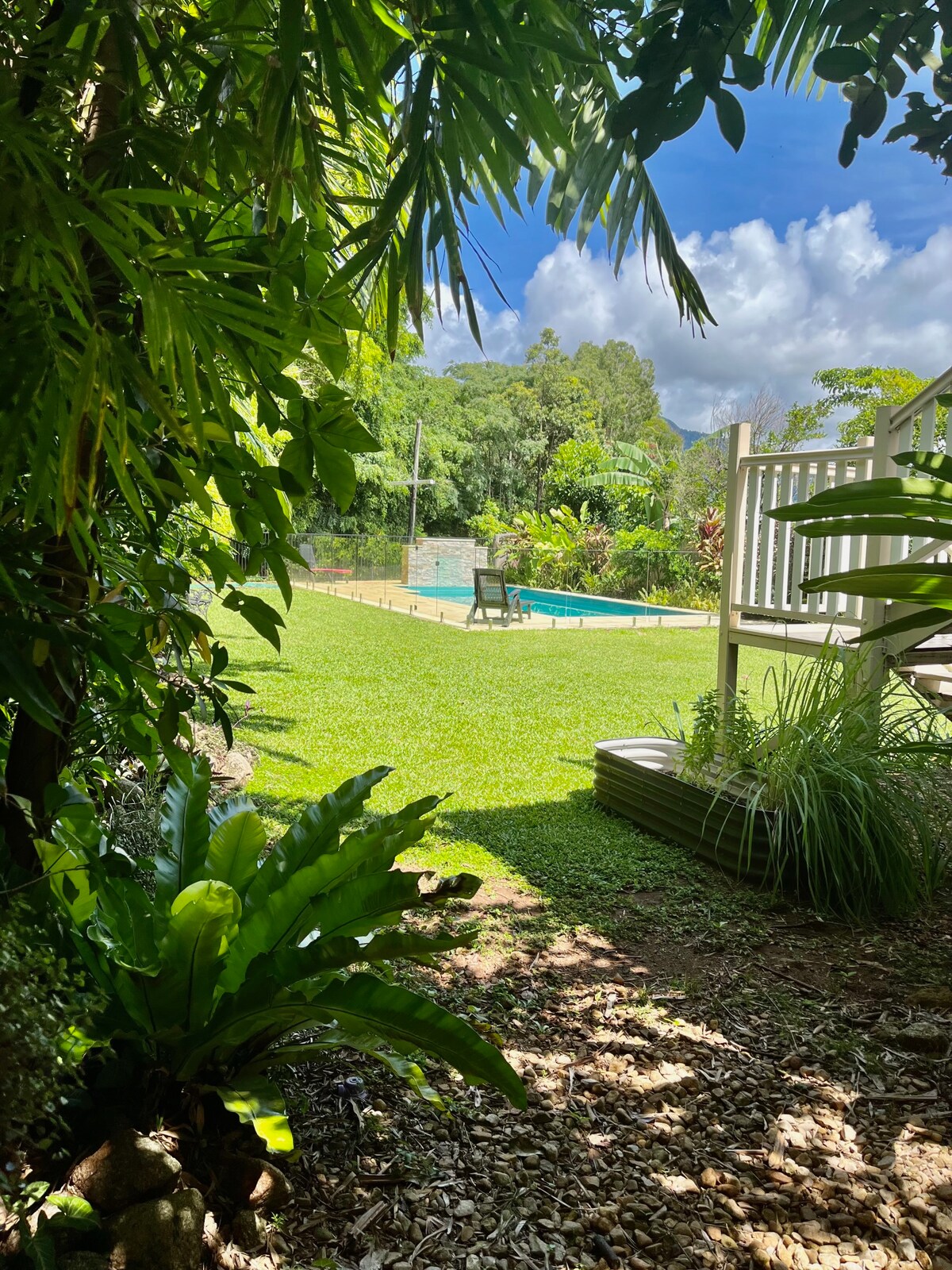 A vibrant garden area is framed by lush greenery, leading to a swimming pool in the distance. The well-maintained lawn is dotted with tropical plants, and a wooden lounger can be seen near the pool. Clear blue skies with scattered clouds add to the inviting outdoor setting.