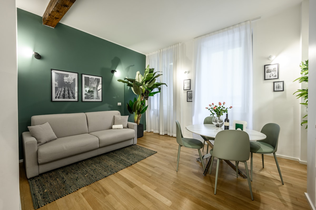 A welcoming living area features a light-colored sofa in front of a vibrant green accent wall. A round dining table with four chairs sits nearby, accompanied by a fresh flower arrangement. Natural light filters through sheer curtains, enhancing the open, airy feel of the space.