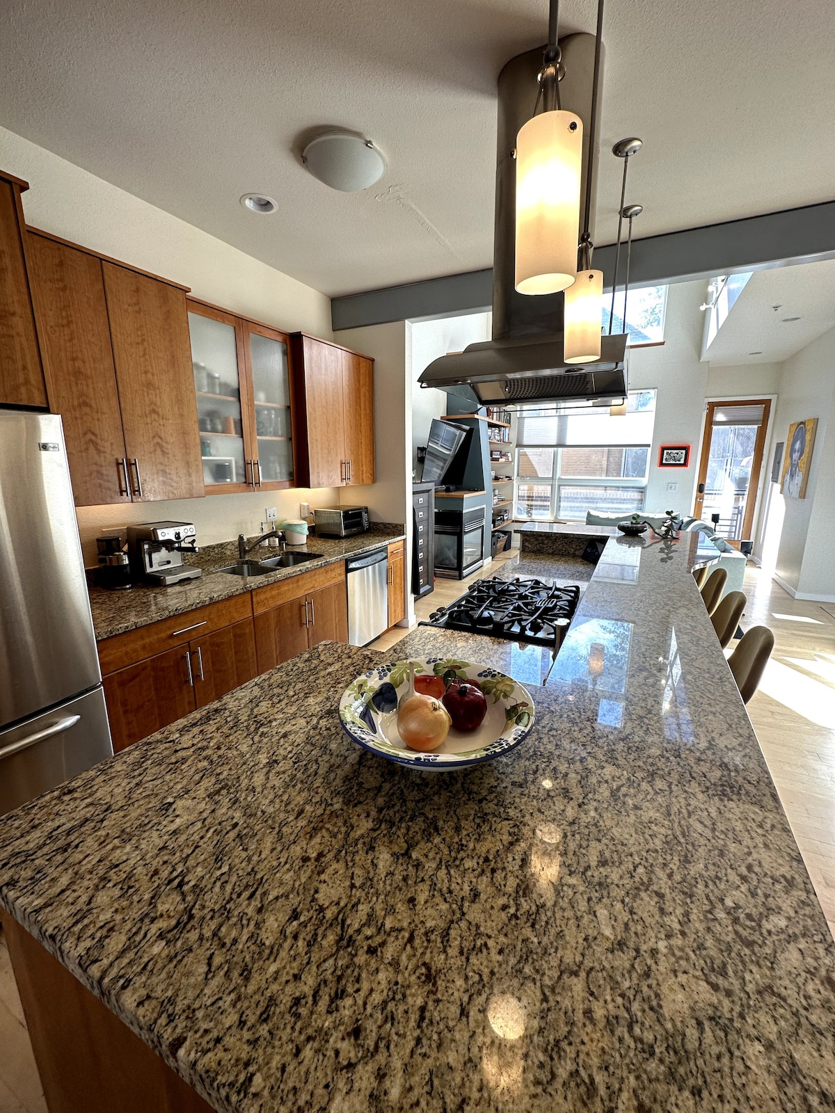 The kitchen features an inviting granite countertop with various fresh fruits displayed in a decorative bowl. Natural light floods the space through tall windows, highlighting the modern stainless-steel appliances and cabinetry. A spacious layout accommodates bar seating for casual dining.