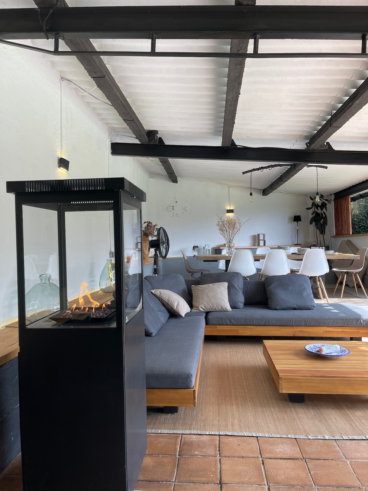 A modern living area features a cozy sofa arrangement in neutral tones, complemented by a sleek glass fireplace. A wooden coffee table sits in front, while a dining area with white chairs and a wooden table is visible in the background, illuminated by soft overhead lighting.