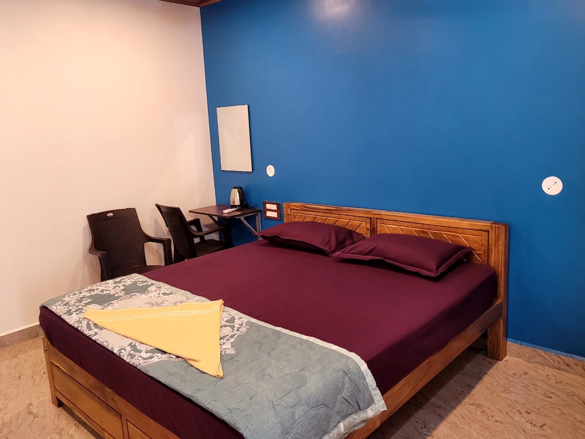 A spacious sleeping area features a large wooden bed with a dark comforter and a light throw. A small desk with a kettle sits nearby. A wall-mounted mirror complements the blue accent wall, while two simple chairs are positioned to the side.
