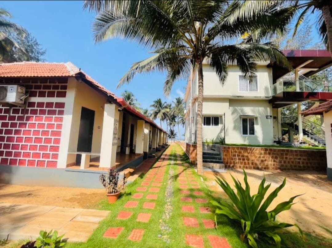 A pathway bordered by green grass and decorative plants is seen, leading between two residential buildings. Palm trees are positioned alongside the structures under a bright blue sky, contributing to a relaxed, tropical setting.