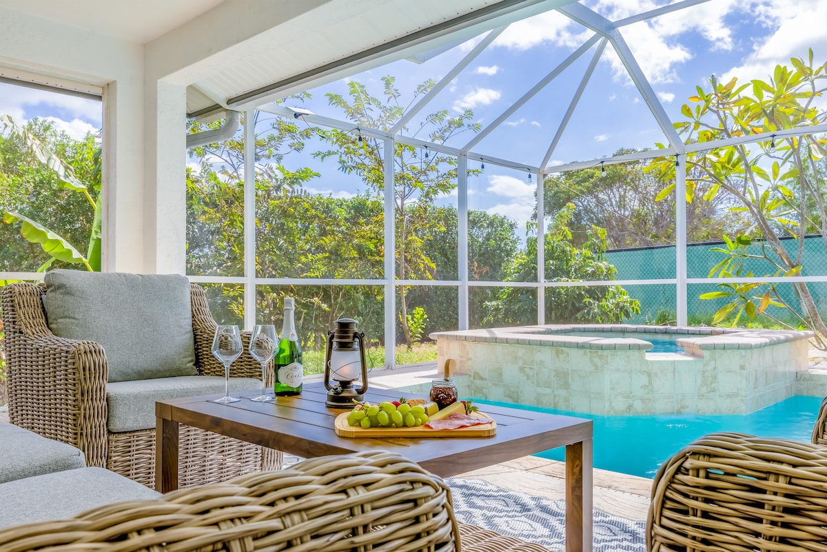 An inviting seating area is presented with wicker chairs and a wooden table, set beside a sparkling pool. A bottle of sparkling wine, two glasses, and a platter of fruit are arranged on the table. Sunlight filters through the screen, highlighting lush greenery beyond.