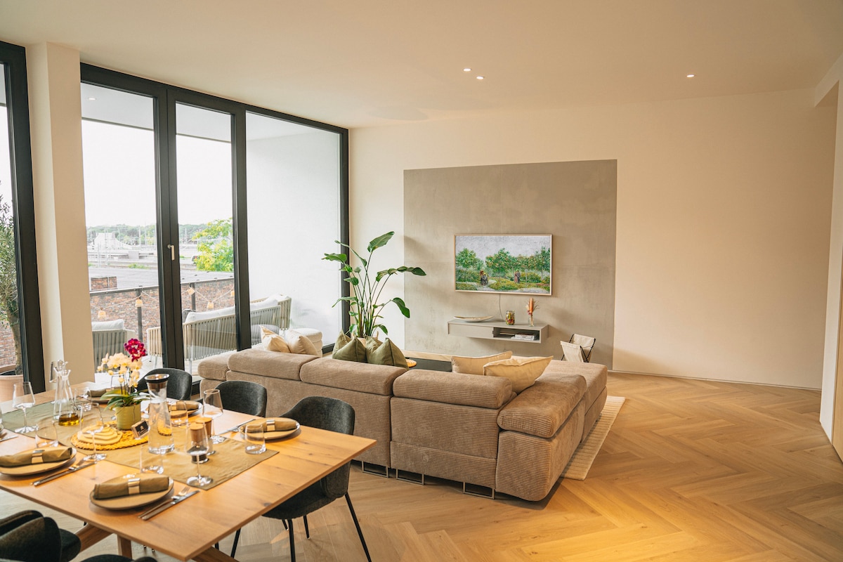 A spacious living area is presented, featuring a large sectional sofa and a minimalist entertainment unit with a mounted television. A dining table is set for meals, while large windows provide natural light and a view of the outdoor terrace.