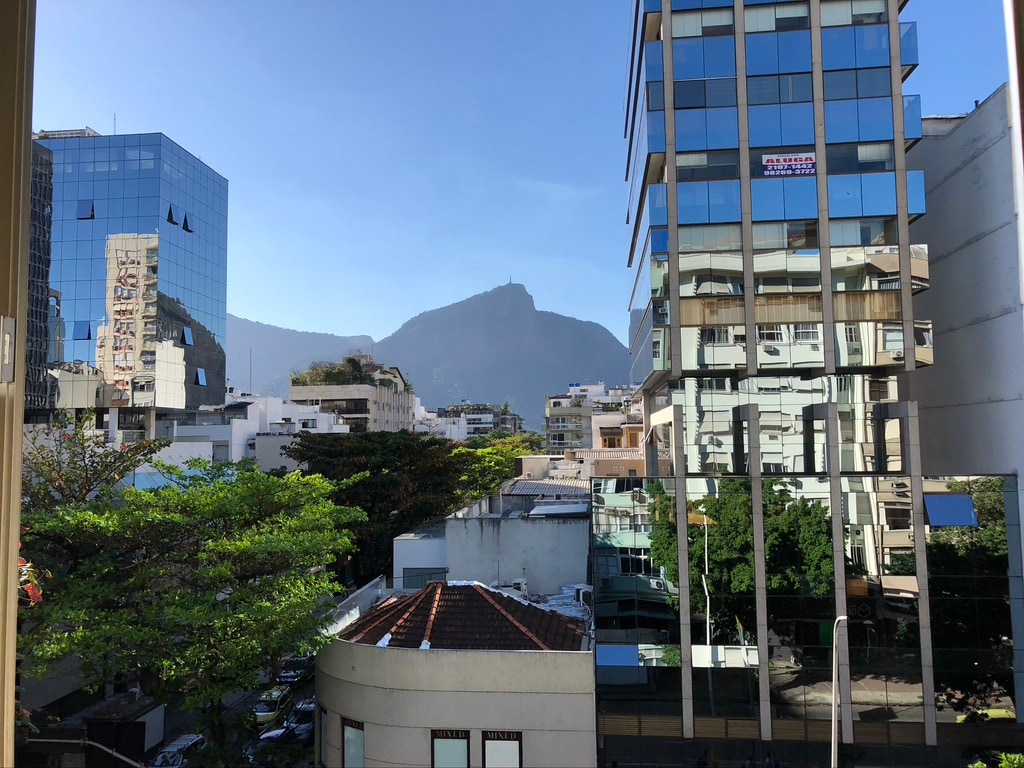 A clear view showcases the Corcovado mountain in the distance, framed by modern buildings. The greenery of nearby trees contrasts with the urban landscape, offering a vibrant backdrop to the scene. Natural light brightens the area, enhancing the appeal of the urban surroundings.