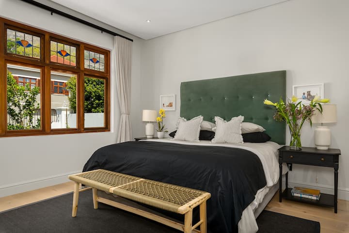 Svetlá, štýlová spálňa s luxusným zeleným čelom, elegantnou výzdobou a vitrážovými oknami pre ďalší šarm. Užite si útulnú atmosféru s mäkkou posteľnou bielizňou, nočnými lampami a bujnou zeleňou za oknom, čo vytvára pokojné útočisko.