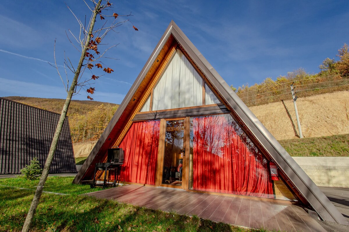 An A-frame cabin features a sloped roof and large windows adorned with vibrant red curtains. The wooden deck extends out to a grassy area, providing a direct connection to the surrounding nature. Trees are visible in the foreground against a clear blue sky.