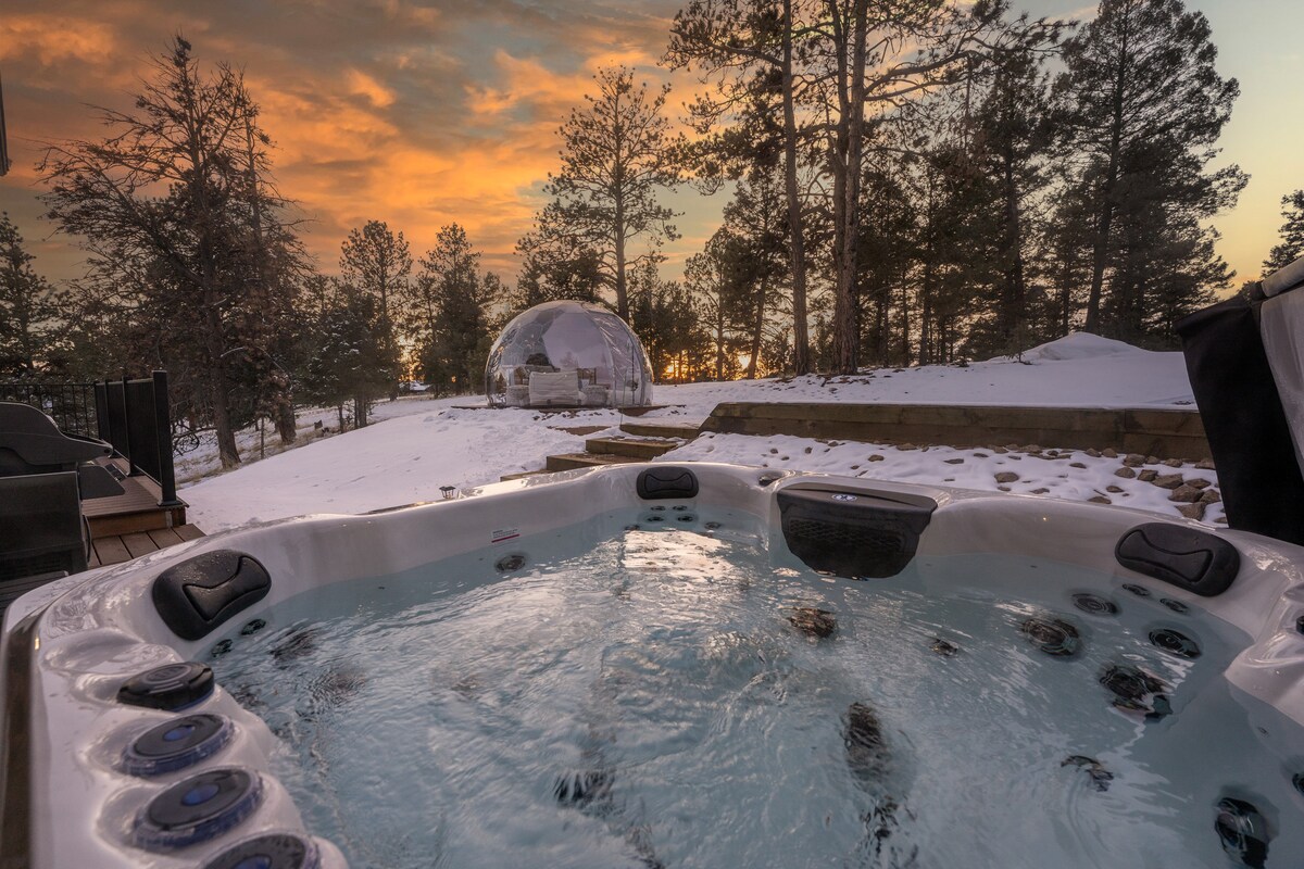 A private hot tub sits bubbling with clear water, surrounded by a snowy landscape. In the background, a geodesic stargazing dome is set against a vibrant sunset sky, framed by towering pine trees creating a serene atmosphere.