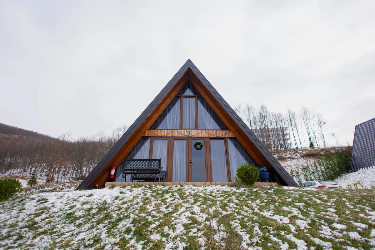 A charming A-frame cabin is set against a serene landscape. Its sloping roofline is complemented by large windows that reflect the surroundings. A front porch contains a simple bench, and a dusting of snow covers the grassy area, enhancing the tranquil ambiance.