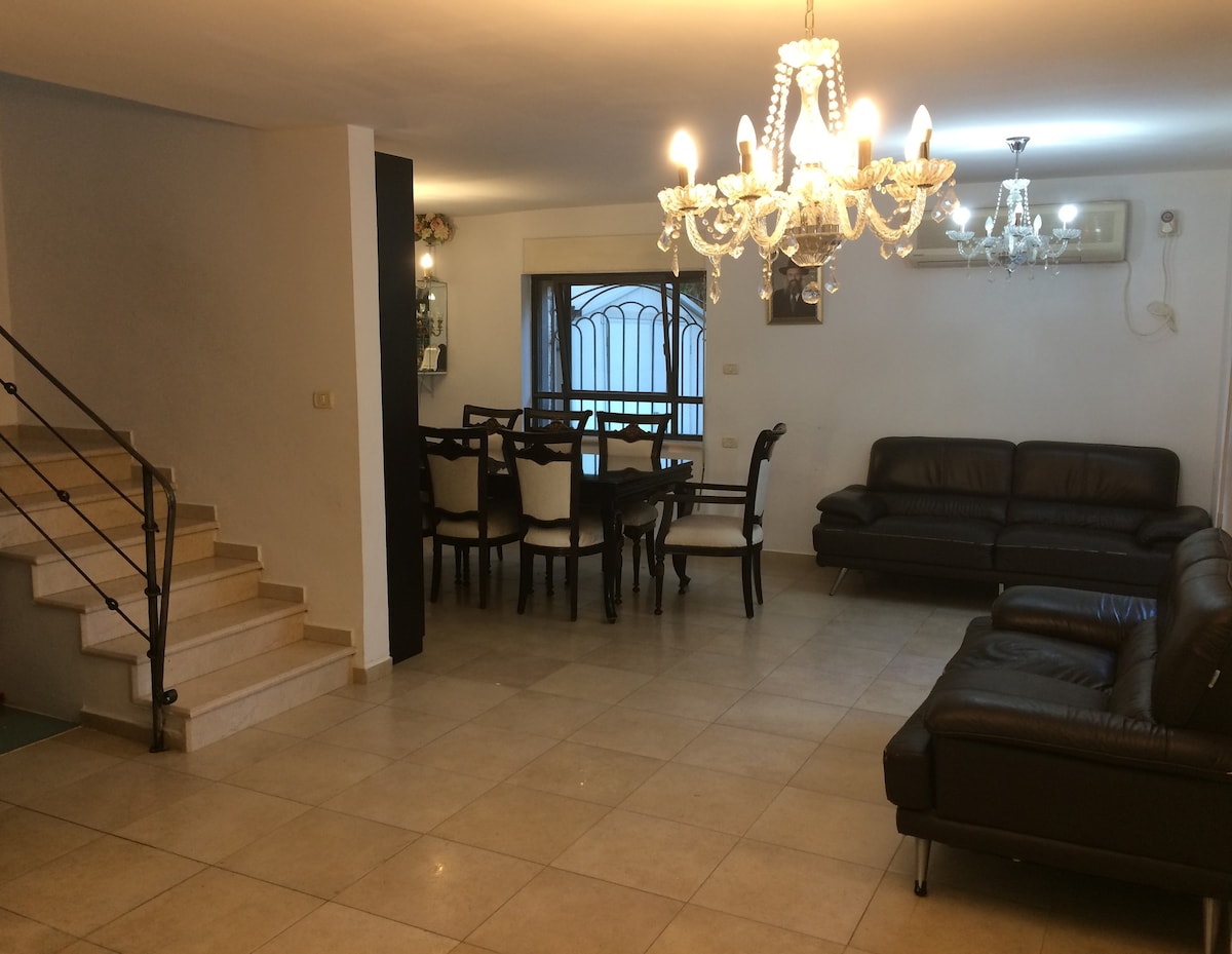 A spacious living area features a modern design, with comfortable dark leather sofas arranged for social gatherings. A large dining table is set for family meals. Bright overhead chandeliers illuminate the space, enhancing the open feel. A staircase is visible in the background.