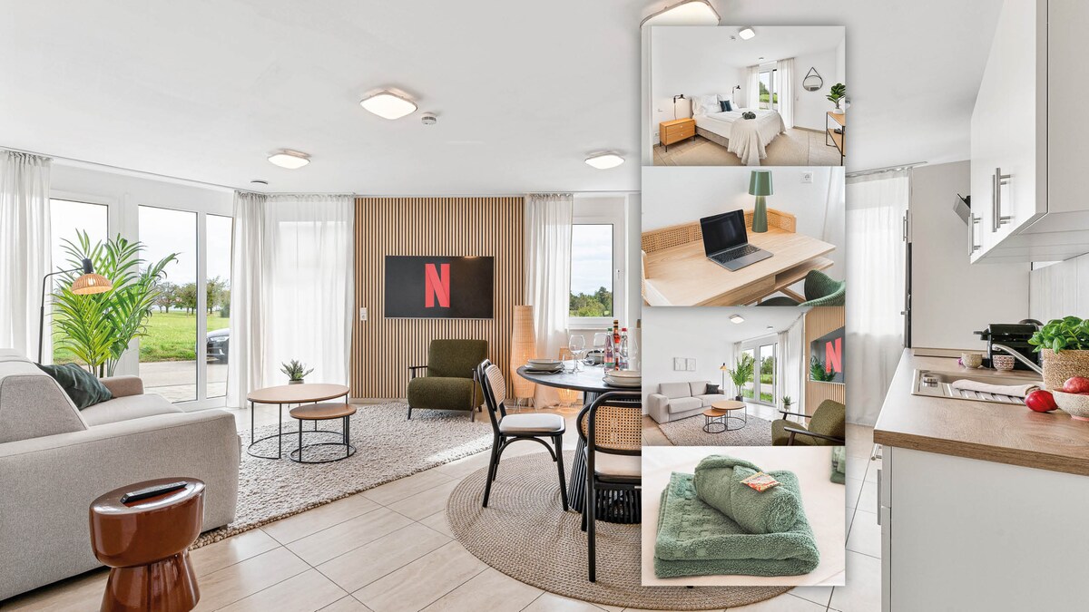 A spacious suite featuring an open layout with a cozy seating area, dining space, and a modern kitchen. Large windows allow natural light to fill the room, while a Smart-TV is mounted on the wall. A subtle decor includes plants and contemporary furniture set against a neutral palette.