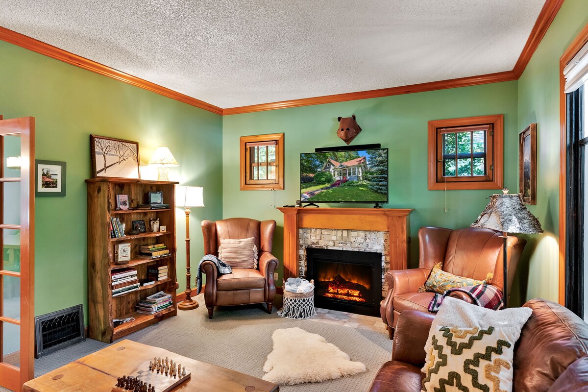 A cozy family room features green walls and wooden accents, highlighting a stone fireplace beneath a wall-mounted television. Two leather armchairs are positioned for comfort, with a small bookshelf nearby. Natural light enters through multiple windows, creating a warm and welcoming environment.