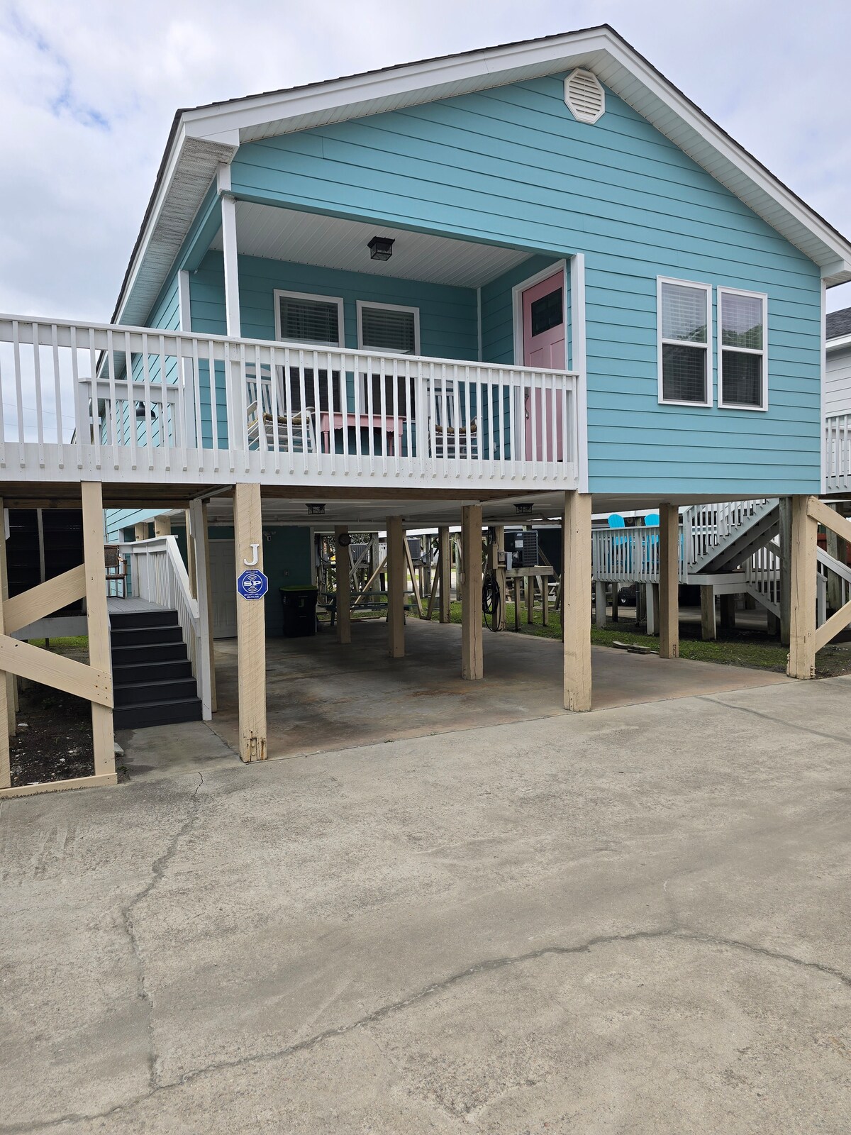 A charming cottage is elevated on wooden supports, showcasing a light blue exterior with a welcoming front entrance. The covered porch features white railings, and a driveway leads to ample parking space beneath the cottage.