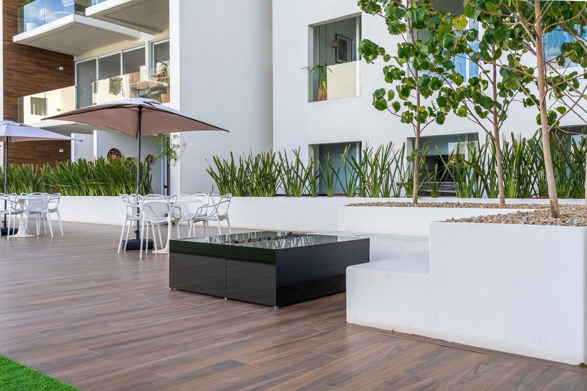 An outdoor area showcases a sleek black firepit surrounded by light-colored seating and tables. Umbrellas provide shade, while lush greenery lines the space, enhancing the relaxed atmosphere. The flooring features warm wooden tones, contrasting with the modern architecture of the building.