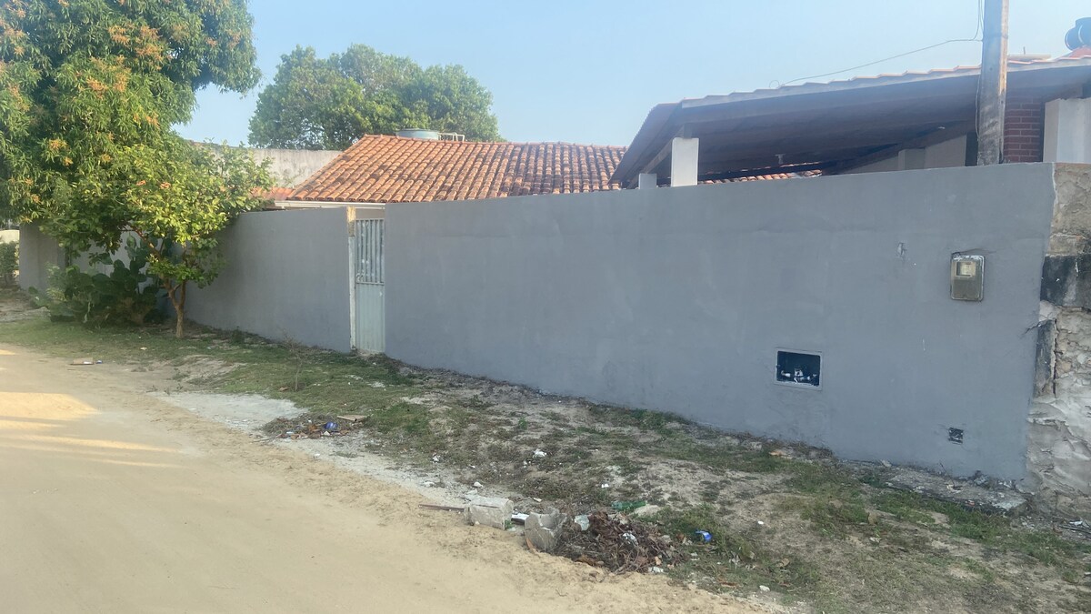 A simple gray wall surrounds a property, partially obscuring a terracotta-tiled roof visible in the background. A narrow gate is positioned along the fence, set against a backdrop of greenery. The ground is sandy with scattered debris, suggesting an untamed outdoor environment.