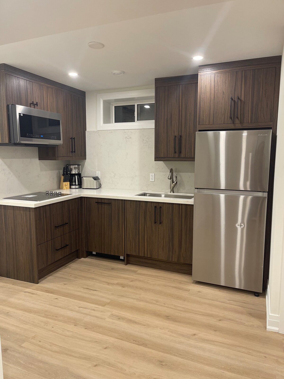 A modern kitchen is shown, featuring brown wooden cabinets and stainless steel appliances. A sleek refrigerator stands beside a compact cooking area, with a double sink and a microwave above. Soft overhead lights illuminate the clean countertops and contemporary design.