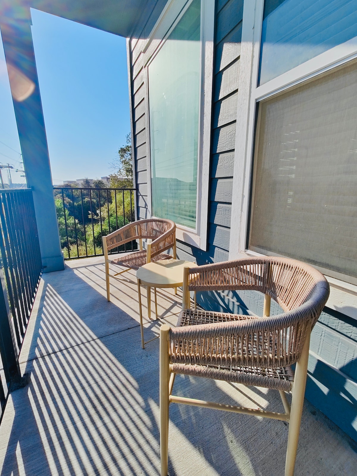 A small balcony is depicted with two woven chairs and a small round table, positioned against the backdrop of large windows. Soft sunlight casts shadows on the concrete floor, enhancing the outdoor space suitable for relaxation.