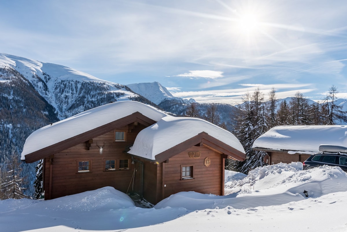 A charming wooden chalet is situated in a snow-covered landscape, surrounded by tall trees and distant mountains. Bright sunlight illuminates the scene, creating a serene winter atmosphere. The structure features a classic sloped roof and a welcoming facade that reflects the authentic Swiss style.
