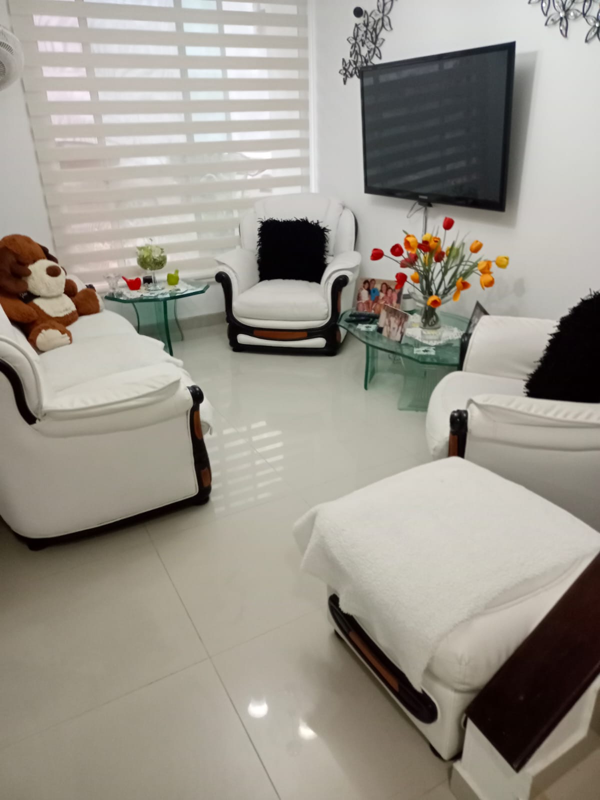 A bright living area features white upholstered chairs complemented by black accent pillows. A plush teddy bear sits on one chair, while a glass table displays a vase of colorful tulips. Natural light filters through sheer blinds, illuminating the sleek tiled floor.