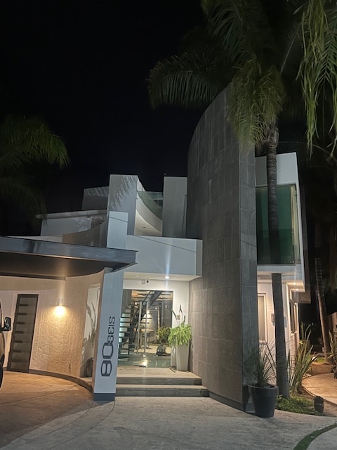 The modern building is illuminated at night, showcasing a sleek facade with a curved design and a mix of stone and glass elements. Decorative plants are positioned at the entrance, and a spacious outdoor area is visible, framed by palm trees.