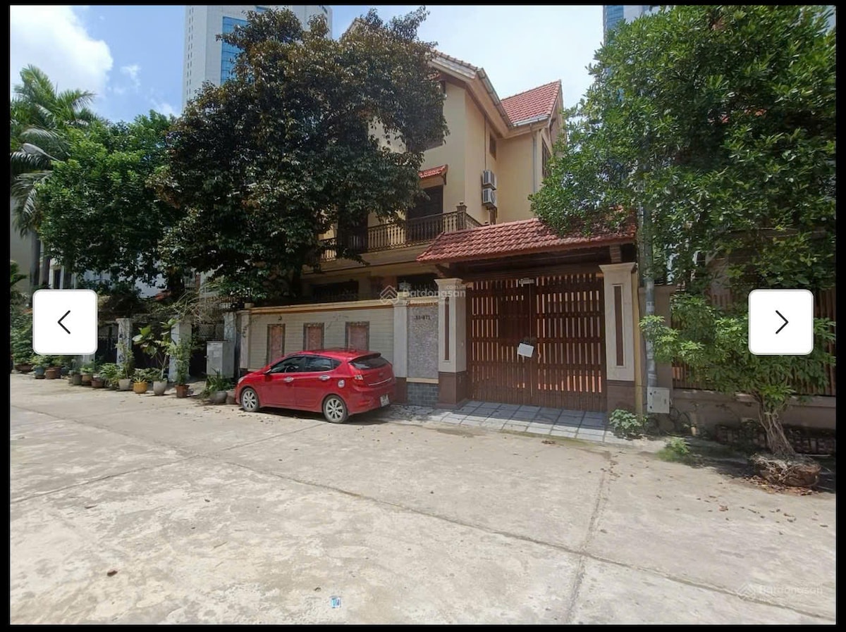 The exterior of the villa is showcased, featuring a two-story structure with a red-tiled roof and decorative wooden elements. A red car is parked nearby, and lush greenery surrounds the property, contributing to a private atmosphere. The entrance is marked by a gated entryway.