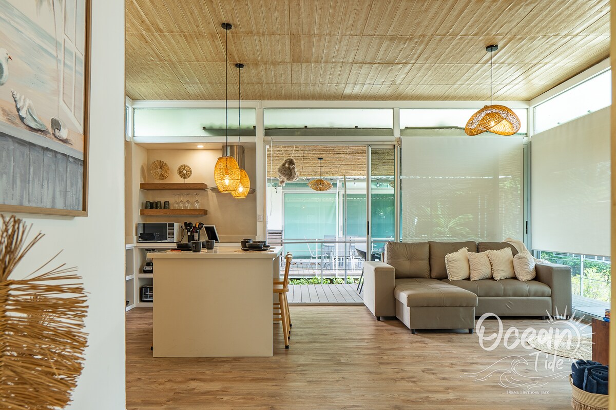 An open living area features a modern kitchen with a seating area, accented by pendant lighting. A spacious couch is positioned to invite relaxation, while large glass doors provide natural light and views of the deck outside.