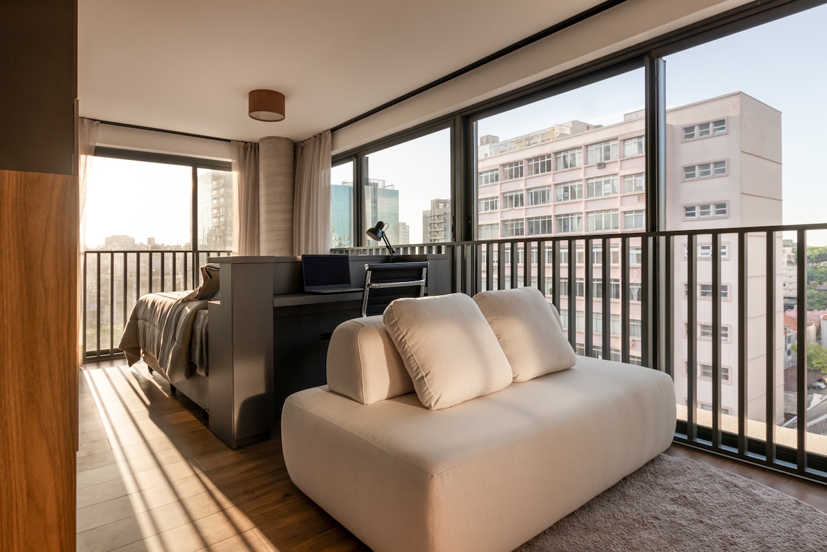 A spacious area is shown featuring large windows that allow natural light to fill the room. A cozy seating arrangement is positioned near the light, while a soft bed is visible in the background, providing a modern yet comfortable aesthetic.