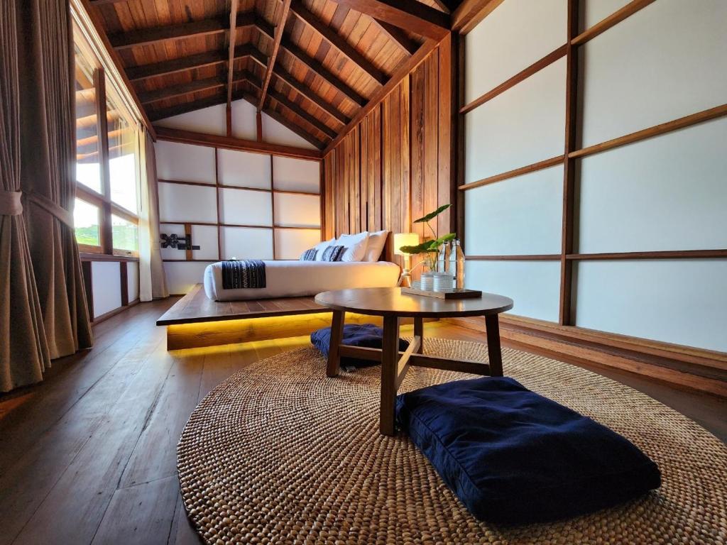 A serene room features wooden walls and a sloped ceiling, illuminated by natural light from large windows. A spacious couch is positioned on a raised platform, accompanied by a round table. Mats are laid on the floor, enhancing the inviting space.