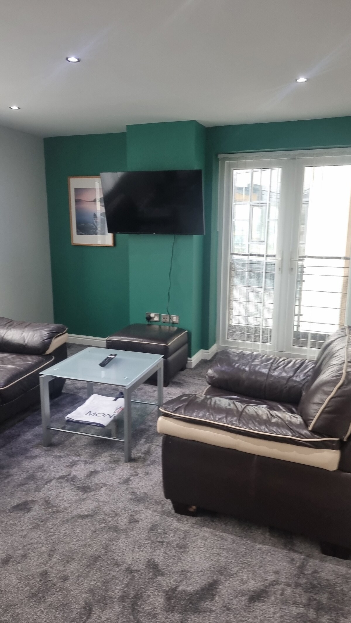A comfortable living area features two dark leather sofas arranged around a glass coffee table. A television is mounted on a green wall, and light enters through a set of glass doors that lead to a balcony.