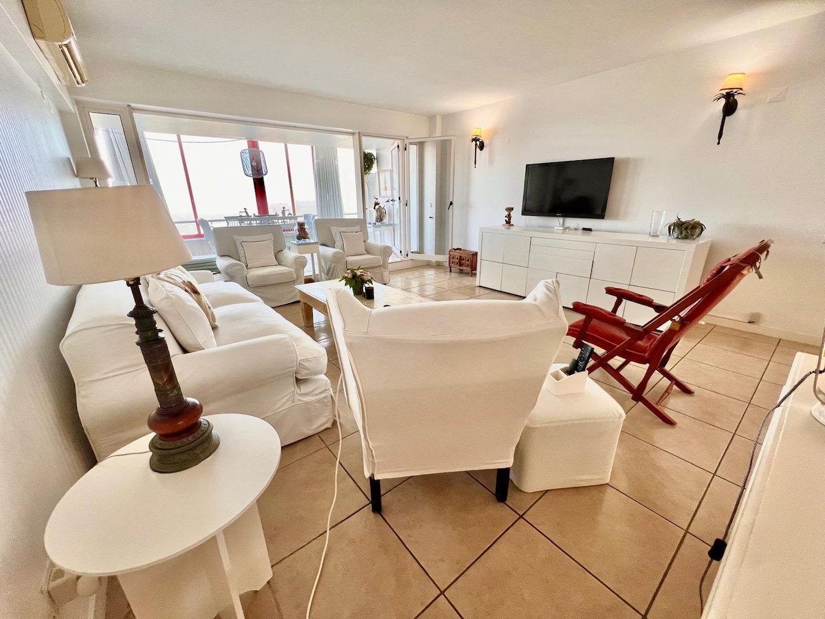 A light-filled living area features comfortable white sofas and armchairs arranged around a small side table. A large flat-screen TV is mounted on a white media unit. Sunlight softly illuminates the tiled floor, highlighting the room's spacious and inviting atmosphere.
