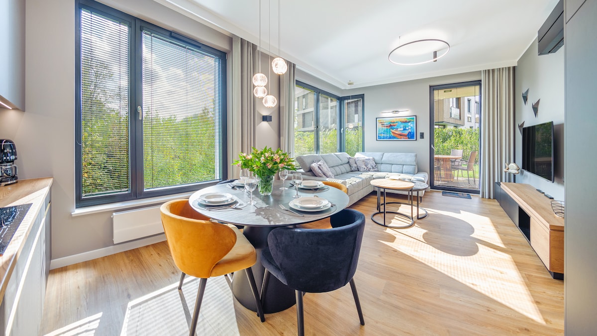 A bright living area features large windows that provide natural light and views of greenery. A modern round dining table is set with tableware, surrounded by plush chairs. A comfortable sofa invites relaxation, complemented by a flat-screen television and a stylish coffee table.
