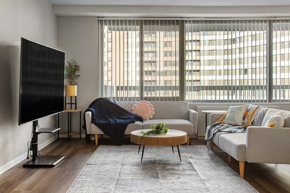A bright living area features two comfortable sofas positioned around a round coffee table. The space is illuminated by natural light streaming through large windows adorned with vertical blinds. A television is mounted on the wall, and a cozy throw blanket is draped over one sofa.