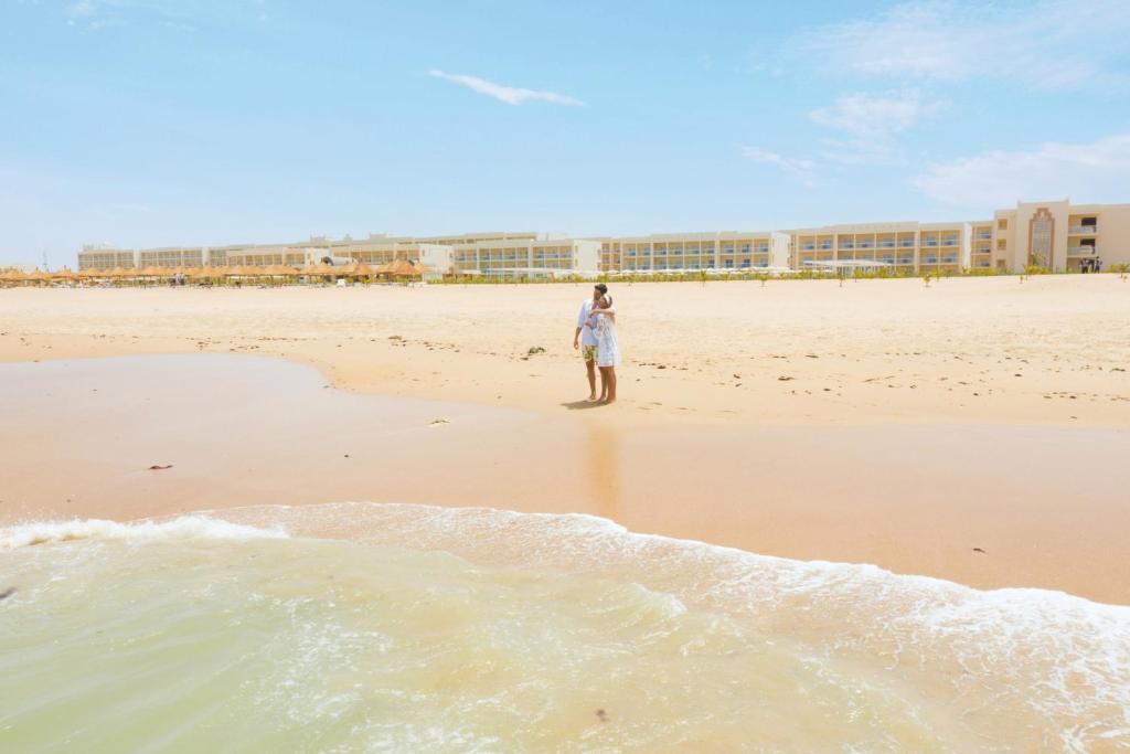 The image shows a couple standing on a sandy beach near the shoreline, with gentle waves lapping at their feet. In the background, the hotel is visible, characterized by modern, elongated buildings against a clear blue sky.
