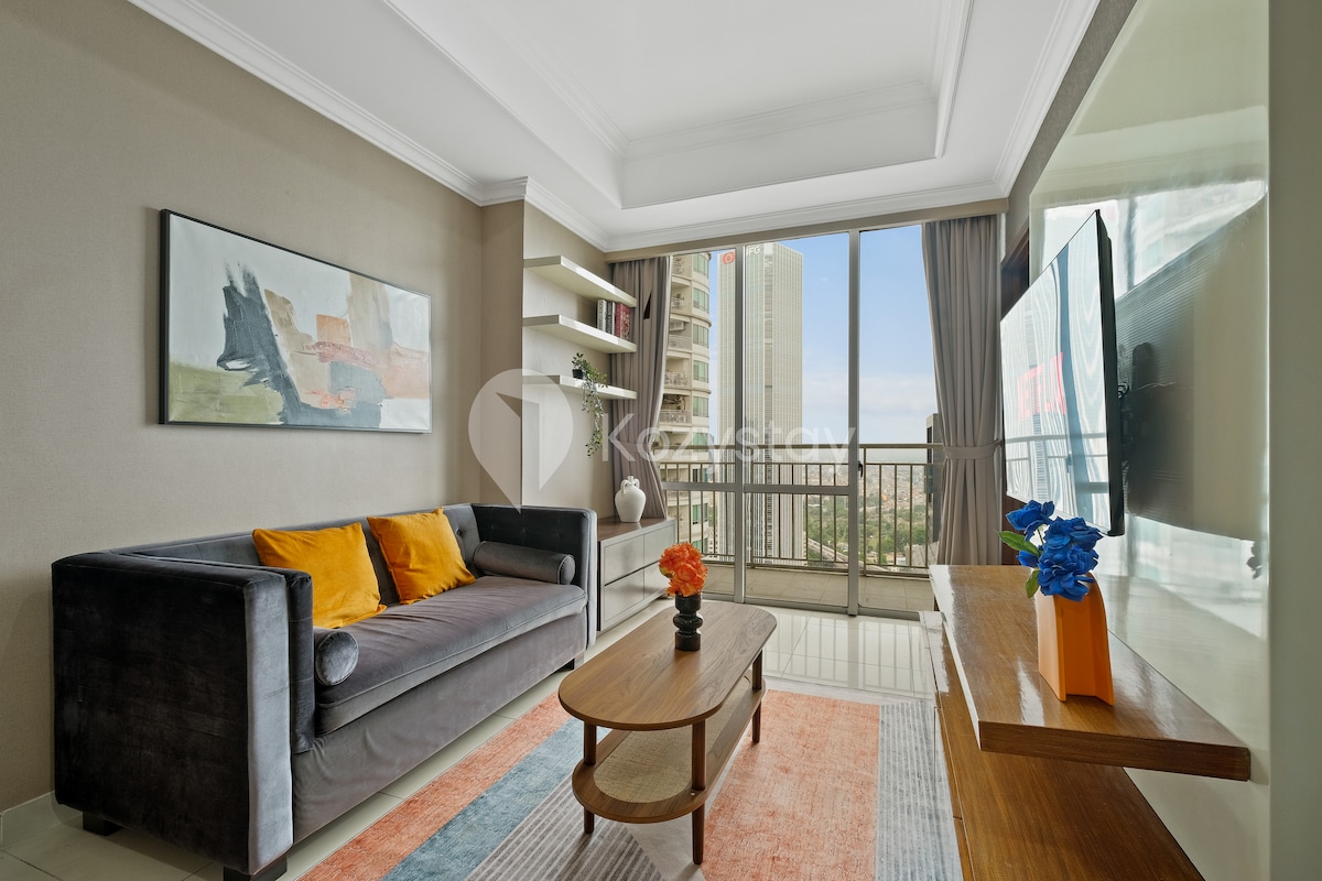 A cozy living area is furnished with a comfortable couch adorned with orange cushions. A wooden coffee table sits on a patterned rug, while a flat-screen TV is mounted on the wall. Large windows offer natural light and a view of the balcony and city skyline.