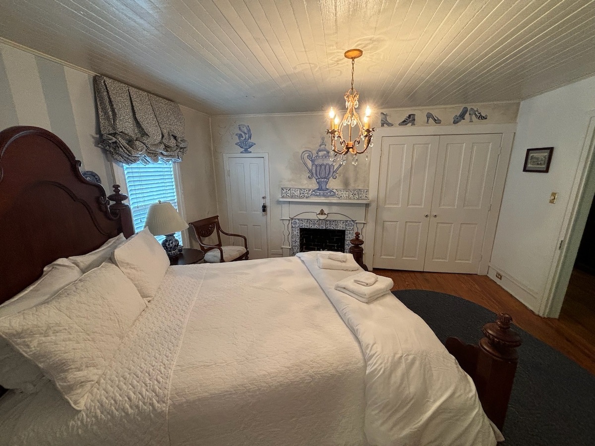 A spacious bedroom features a large bed with white linens, complemented by decorative pillows. A chandelier hangs from the ceiling, providing soft lighting. Additional elements include a vintage chair and a fireplace with a decorative mantel. Light-colored walls are adorned with subtle artwork.