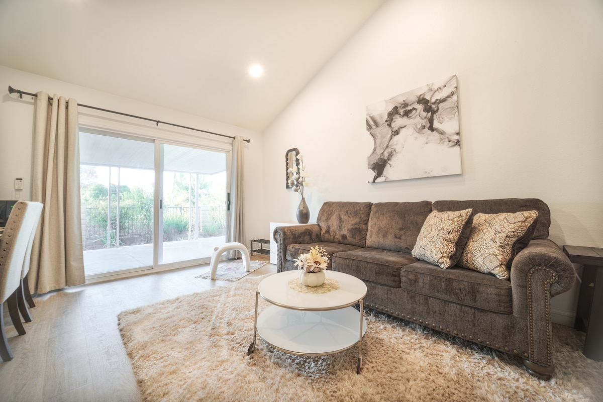 A cozy living area is highlighted by a dark upholstered couch adorned with accent pillows. A circular coffee table sits in front, topped with decorative items. Large sliding glass doors lead to an outdoor space, while neutral-toned curtains frame the windows.