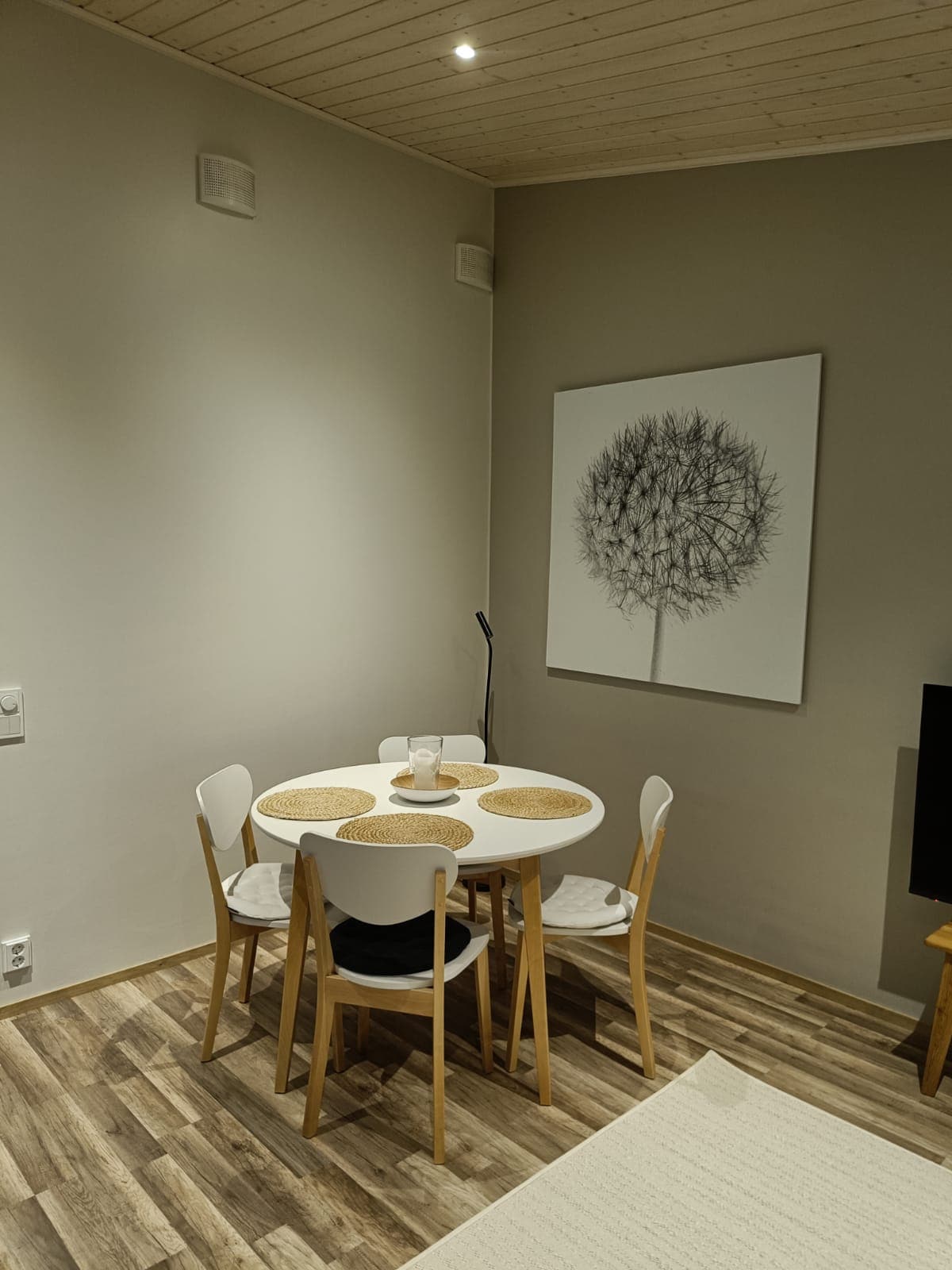 A dining area is present, featuring a round table surrounded by four light-colored chairs. A simple artwork depicting a tree is displayed on the wall, while a floor lamp stands beside the table, providing soft lighting.