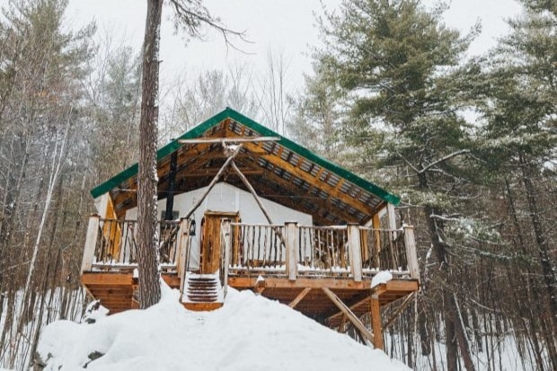 A cabin elevated on stilts is surrounded by snow-covered ground, with a sloped roof covered in green accents. A spacious deck made of wooden planks features railings crafted from logs, providing a serene view of the snowy forest landscape.
