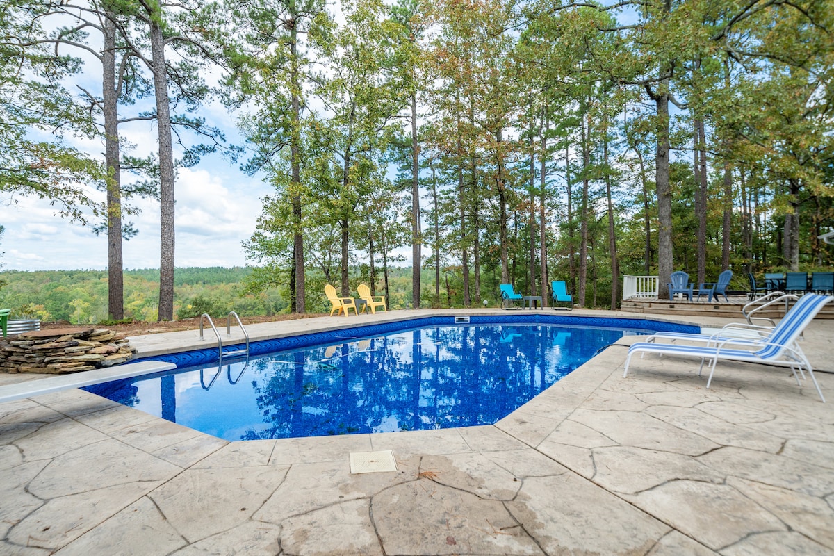 An outdoor pool area is surrounded by a stone patio with lounge chairs positioned nearby. Colorful seating is visible in the background, framed by tall trees that provide a serene, natural setting. The clear blue water reflects the sky, enhancing the peaceful ambiance.